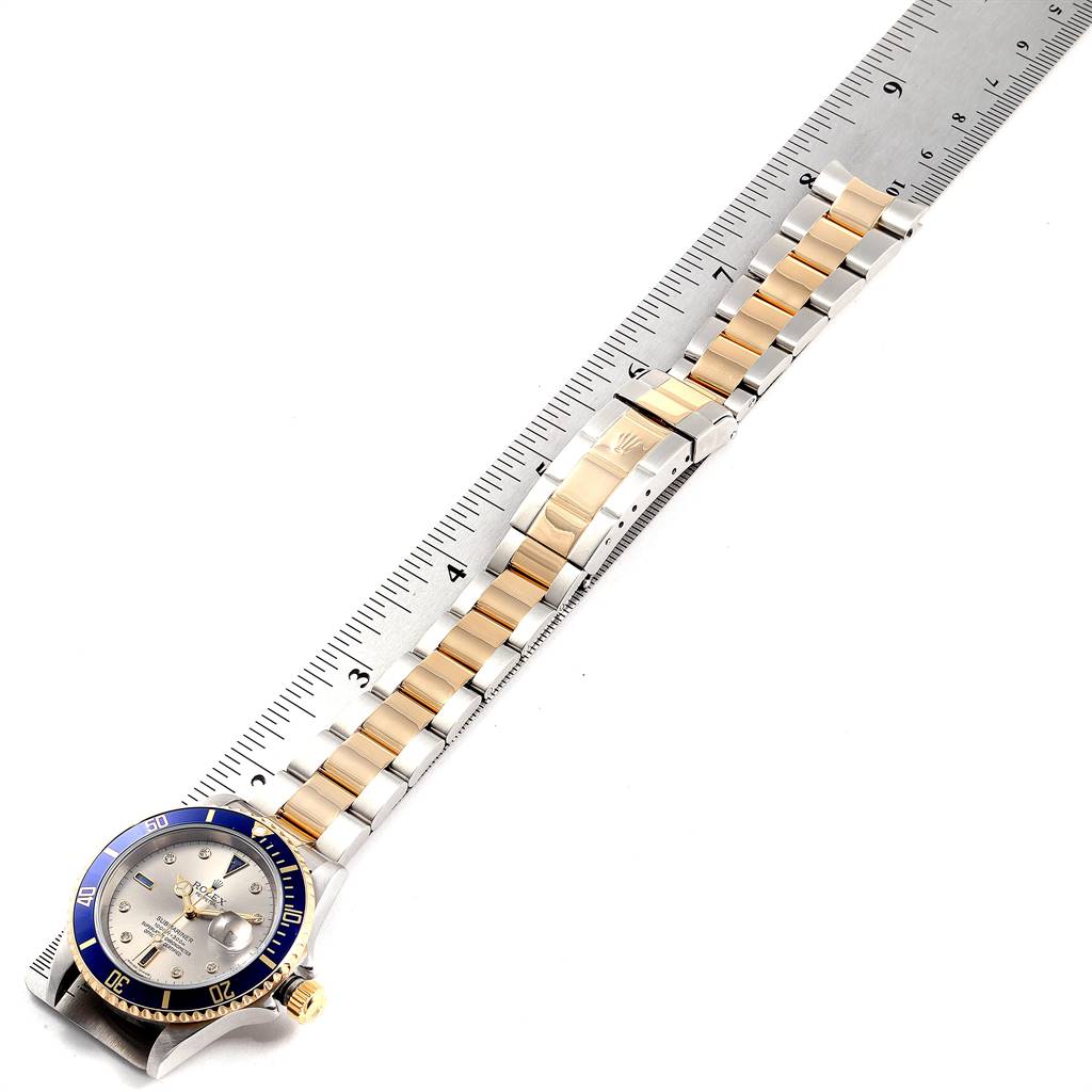 The image shows an overhead view of a Rolex Submariner 16613 Men
s Steel and Gold (two tone) Grey Dial 16613 Men
s Steel and Gold (two tone) Grey Dial watch with a blue bezel and a two-tone band next to a ruler for measurement.