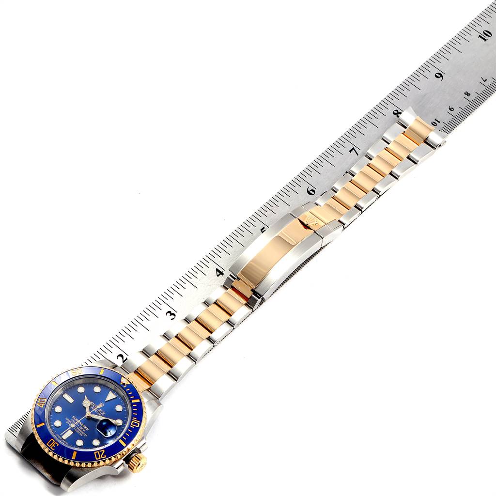 The image shows a Rolex Submariner 116613 Men
s Steel and Gold (two tone) Blue Dial 116613 Men
s Steel and Gold (two tone) Blue Dial watch with a blue dial and bezel, lying flat next to a ruler, highlighting its strap and clasp.