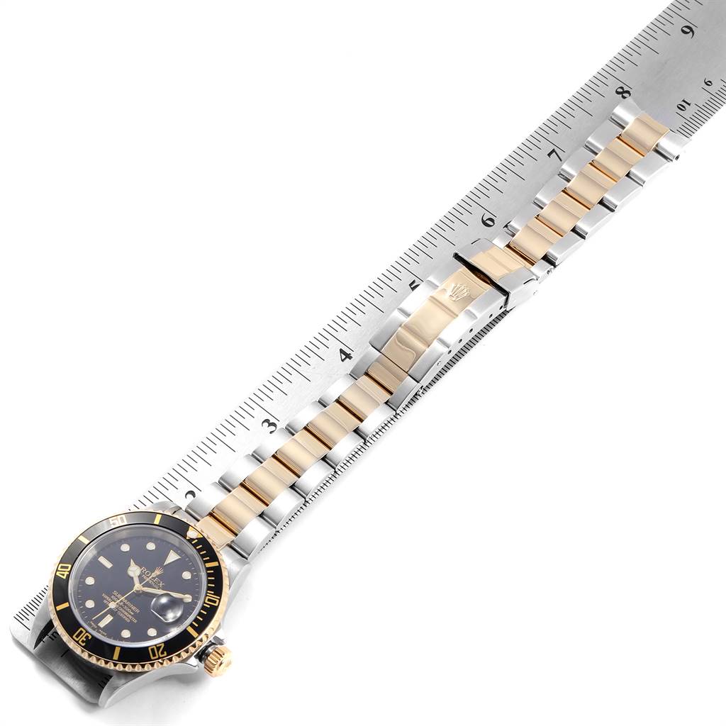 This image shows the Rolex Submariner 16613 Men
s Steel and Gold (two tone) Black Dial 16613 Men
s Steel and Gold (two tone) Black Dial watch laid flat on a ruler, highlighting its gold and silver bracelet and black dial.