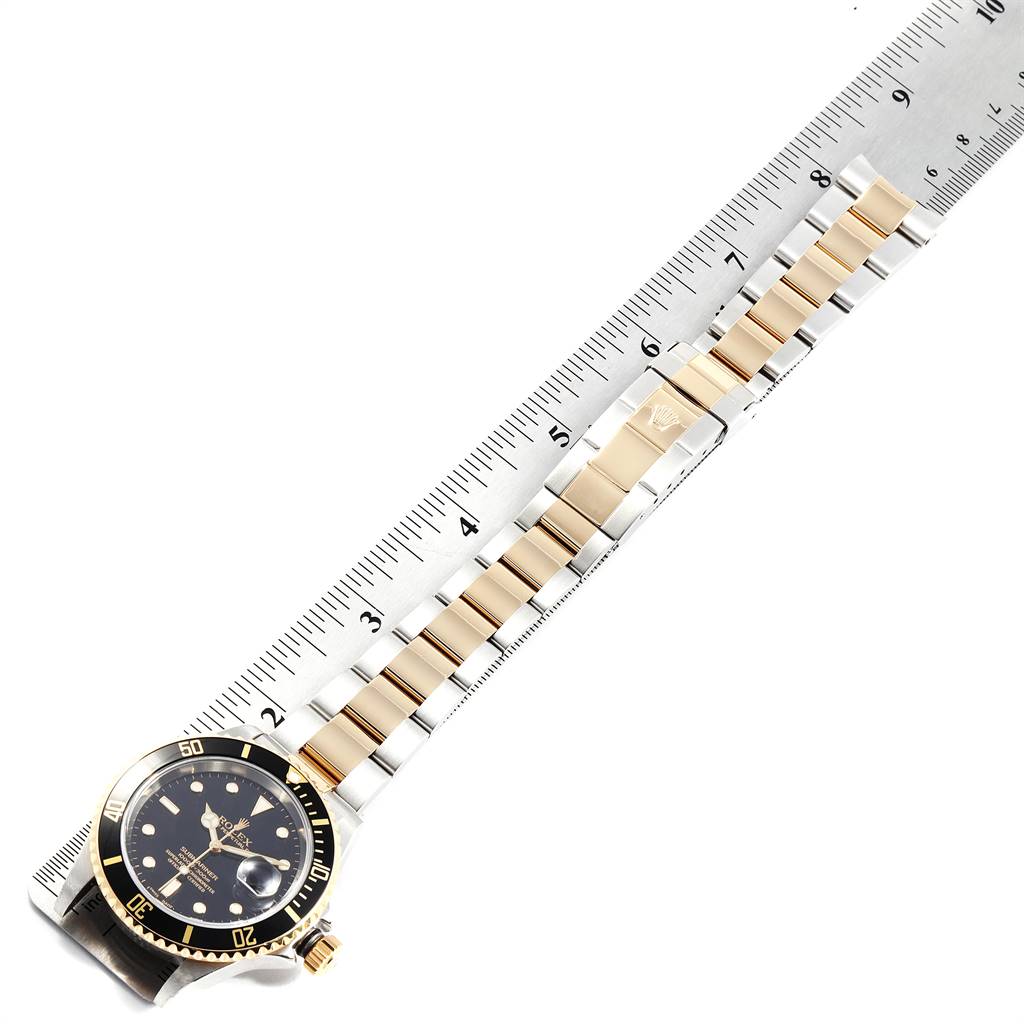 The image shows a Rolex Submariner 16613 Men
s Steel and Gold (two tone) Black Dial 16613 Men
s Steel and Gold (two tone) Black Dial watch laid flat alongside a ruler, highlighting its bracelet and face.