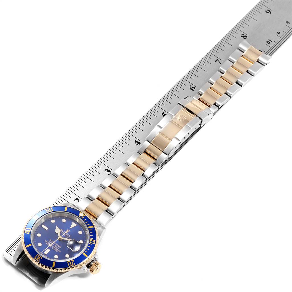 The Rolex Submariner 16613 Men
s Steel and Gold (two tone) Blue Dial 16613 Men
s Steel and Gold (two tone) Blue Dial watch is shown from a top-down angle, displaying its face, bezel, and two-tone bracelet along a ruler.