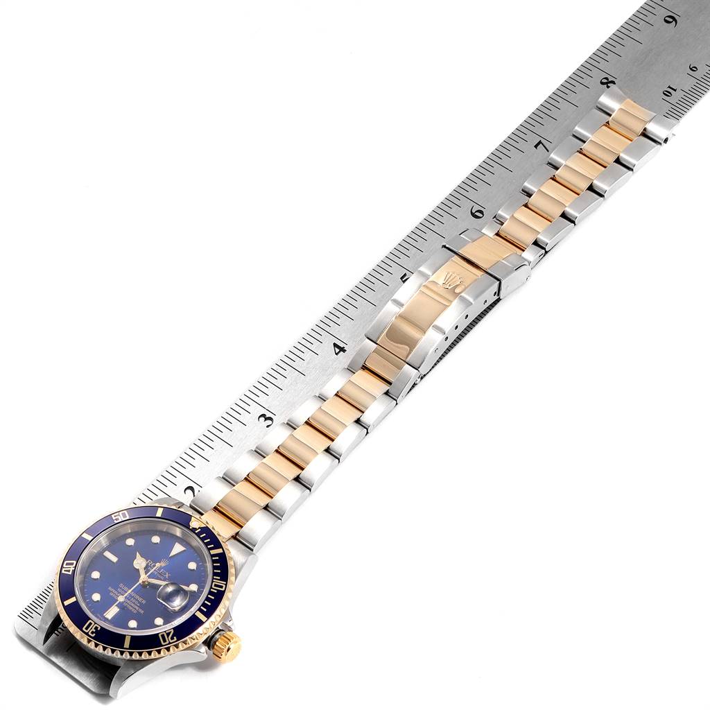 The image shows a top view of a Rolex Submariner 16613 Men
s Steel and Gold (two tone) Blue Dial 16613 Men
s Steel and Gold (two tone) Blue Dial watch with a two-tone metal bracelet placed alongside a ruler.