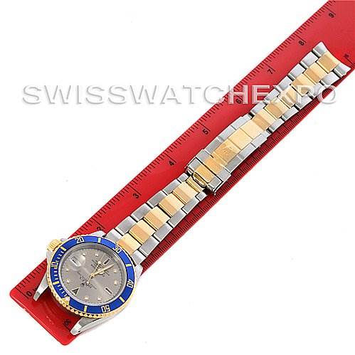 The Rolex Submariner 16613 Men's Yellow Gold Grey Dial 16613 Men's Yellow Gold Grey Dial watch is shown from a top-down angle, displaying its blue and gold bezel, dial, and two-tone bracelet on a red ruler.