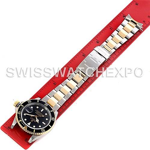 The image shows a top-down view of a Rolex Submariner 16613 Men's Steel and Gold (two tone) Black Dial 16613 Men's Steel and Gold (two tone) Black Dial watch, including its face, bezel, crown, and metal bracelet lying on a red ruler.