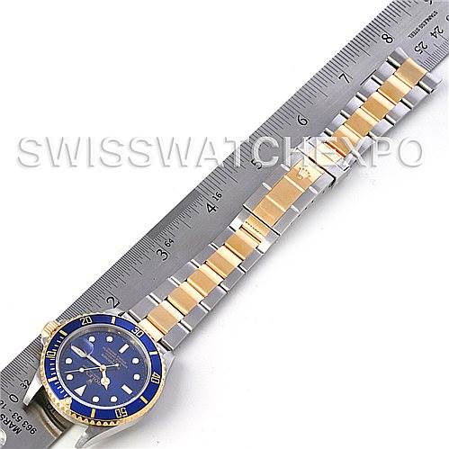 The image shows a Rolex Submariner 16613 Men's Steel and Gold (two tone) Blue Dial 16613 Men's Steel and Gold (two tone) Blue Dial watch laid flat next to a ruler, highlighting its blue dial and two-tone bracelet.