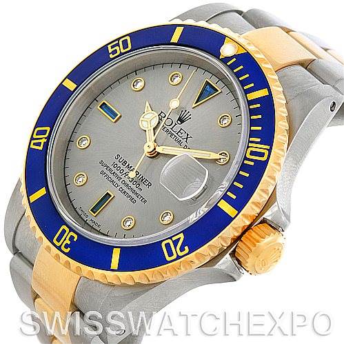 The image shows a Rolex Submariner 16613 Men's Steel and Gold (two tone) Grey Dial 16613 Men's Steel and Gold (two tone) Grey Dial watch, highlighting its blue and gold bezel, grey dial, and metal bracelet from an angled top view.