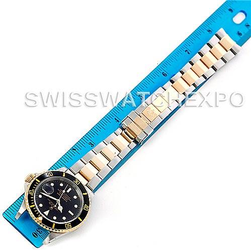 The image shows an overhead view of a Rolex Submariner 16613 Men's Steel and Gold (two tone) Black Dial 16613 Men's Steel and Gold (two tone) Black Dial watch with a two-tone metal bracelet and a blue ruler measuring the length.