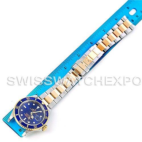 The image shows a top-down view of a Rolex Submariner 16613 Men's Steel and Gold (two tone) Blue Dial 16613 Men's Steel and Gold (two tone) Blue Dial watch with a blue and gold bezel and a metal bracelet placed next to a blue ruler.