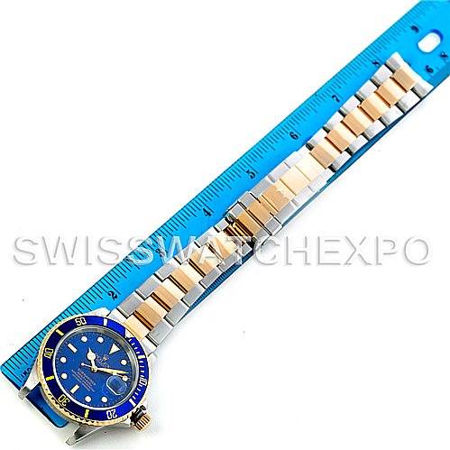 The Rolex Submariner 16613 Men's Steel and Gold (two tone) Blue Dial 16613 Men's Steel and Gold (two tone) Blue Dial watch is shown laid flat next to a blue ruler, highlighting its gold and silver bracelet.