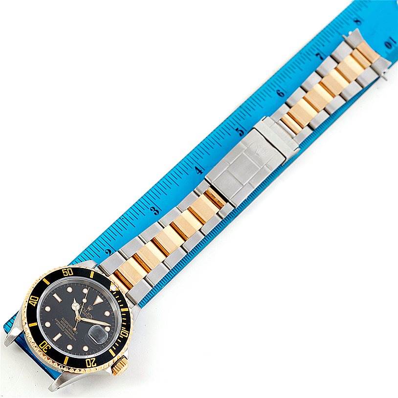 The image shows a top view of a Rolex Submariner 16613 Men's Steel and Gold (two tone) Black Dial 16613 Men's Steel and Gold (two tone) Black Dial watch with a two-tone metal bracelet, placed alongside a blue ruler.