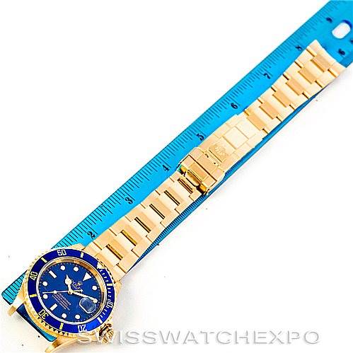 The Rolex Submariner 16618 Men's Yellow Gold Blue Dial 16618 Men's Yellow Gold Blue Dial watch is shown at a top-down angle, featuring its full gold bracelet and blue dial alongside a blue ruler.