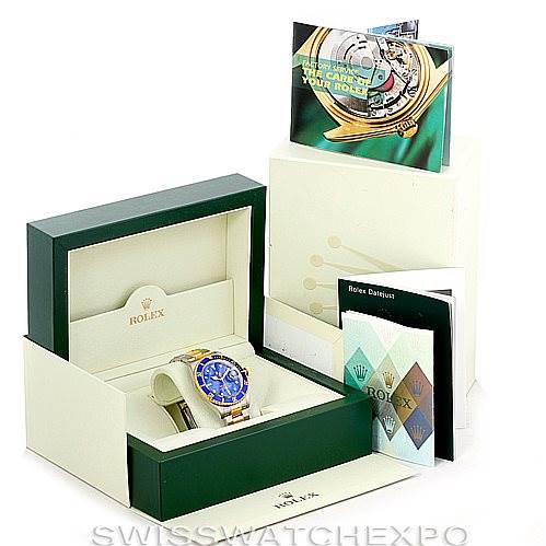 The image shows a Rolex Submariner 16613 Men's Steel and Gold (two tone) Blue Dial 16613 Men's Steel and Gold (two tone) Blue Dial watch angled inside its open box, accompanied by booklets and a green outer packaging.
