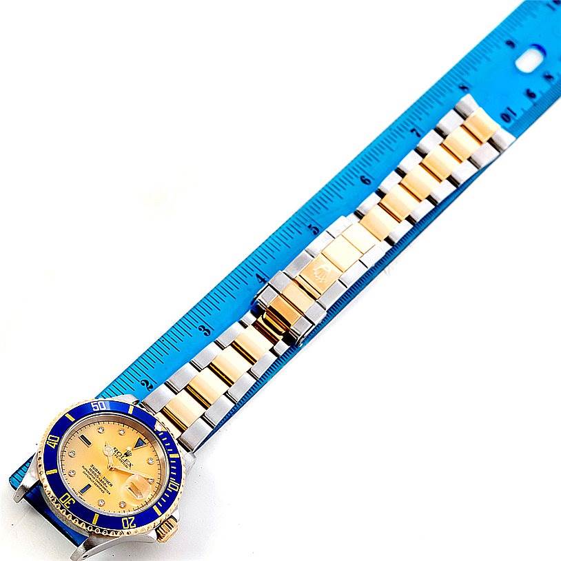 The image shows a Rolex Submariner 16613 Men's Steel and Gold (two tone) Champagne Dial 16613 Men's Steel and Gold (two tone) Champagne Dial watch laid flat next to a blue ruler, displaying both the watch face and the bracelet.