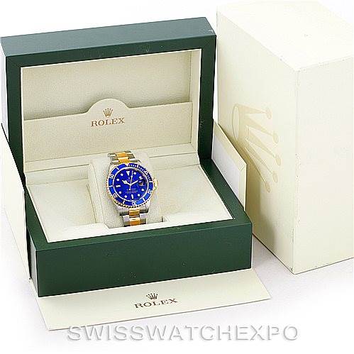 The image shows a Rolex Submariner 16613 Men
s Steel and Gold (two tone) Blue Dial 16613 Men
s Steel and Gold (two tone) Blue Dial watch with a blue dial inside an open green Rolex box, accompanied by outer packaging.