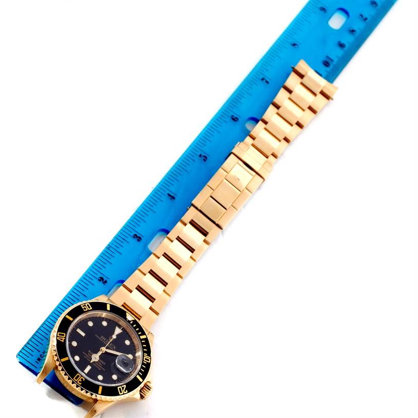 The Rolex Submariner 16618 Men
s Yellow Gold Black Dial 16618 Men
s Yellow Gold Black Dial watch is displayed flat with its gold bracelet extended next to a blue ruler.