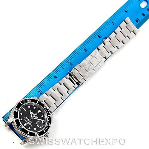 The Rolex Submariner 16610 Men
s Stainless Steel Black Dial 16610 Men
s Stainless Steel Black Dial watch is shown from a top-down angle, displaying the face, bezel, and fully extended bracelet alongside a blue ruler for scale.