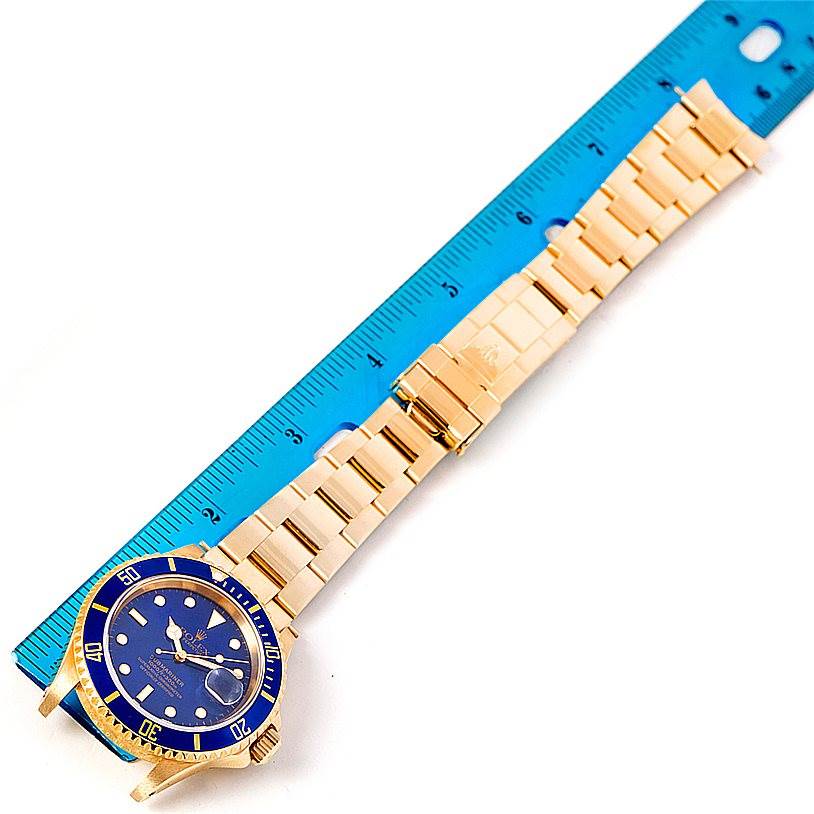 The image shows a top-down view of a Rolex Submariner 16618 Men
s Yellow Gold Blue Dial 16618 Men
s Yellow Gold Blue Dial watch with a gold bracelet next to a blue ruler.