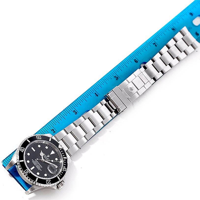 The image shows a Rolex Submariner 16610 Men
s Stainless Steel Black Dial 16610 Men
s Stainless Steel Black Dial watch and its metal strap laid next to a blue ruler.