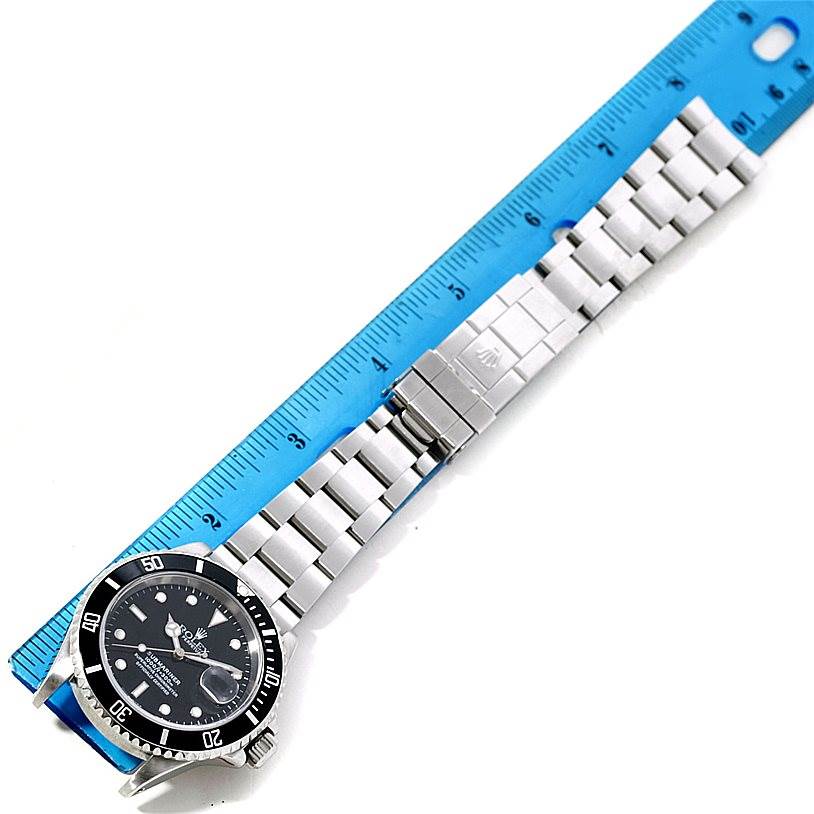 The Rolex Submariner 16610 Men
s Stainless Steel Black Dial 16610 Men
s Stainless Steel Black Dial watch is shown from an overhead angle, displaying its face, bezel, and stainless steel bracelet next to a blue ruler.