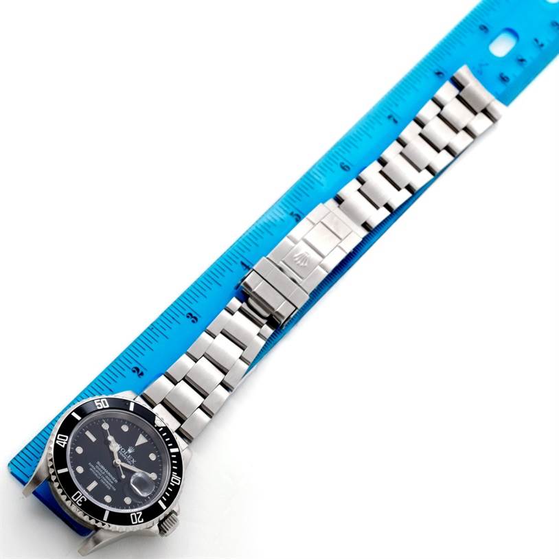 This image shows a Rolex Submariner 16610 Men
s Stainless Steel Black Dial 16610 Men
s Stainless Steel Black Dial watch and its stainless steel bracelet laid flat next to a blue ruler.