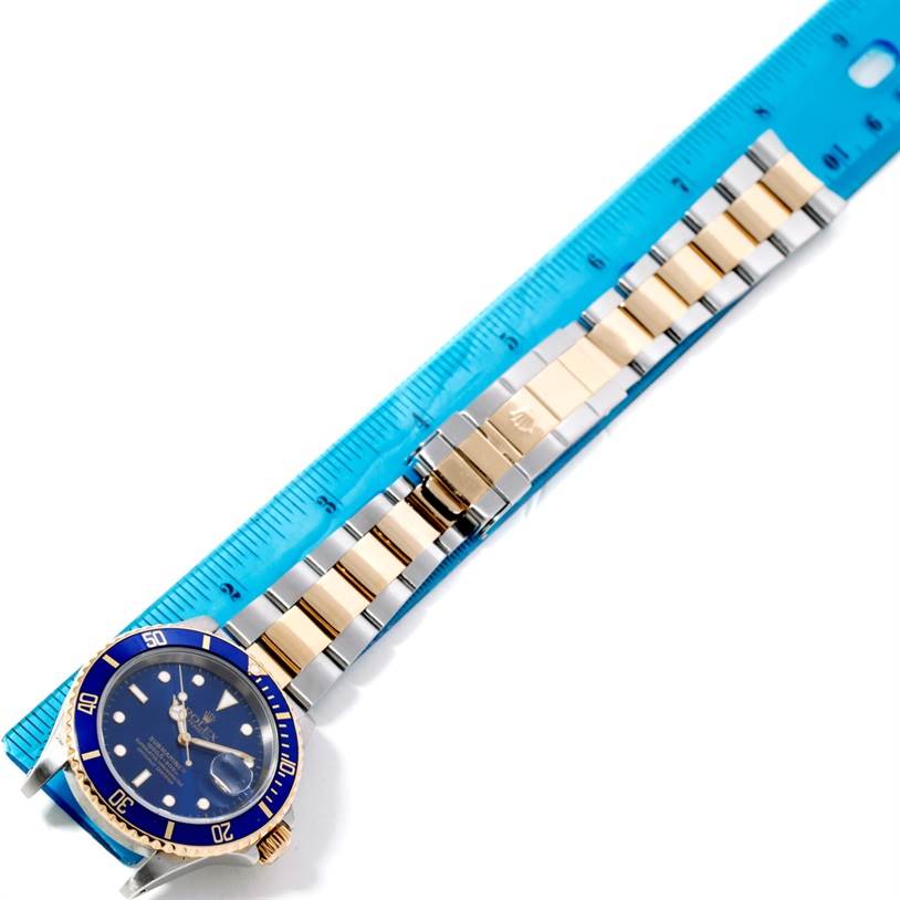 The image shows a Rolex Submariner 16613 Men's Steel and Gold (two tone) Blue Dial 16613 Men's Steel and Gold (two tone) Blue Dial watch with a blue dial and bezel, alongside a ruler for scale.