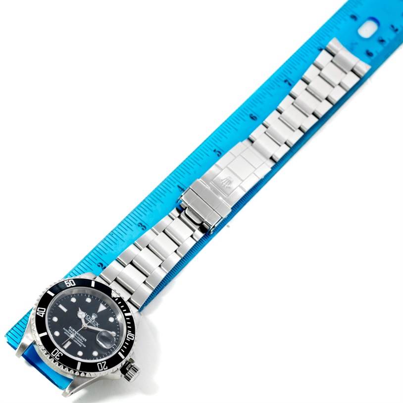 The image shows a Rolex Submariner 16610 Men
s Stainless Steel Black Dial 16610 Men
s Stainless Steel Black Dial watch laid flat with its stainless steel bracelet open, next to a blue measuring ruler.