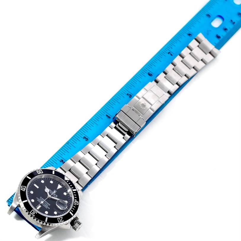 The Rolex Submariner 16610 Men
s Stainless Steel Black Dial 16610 Men
s Stainless Steel Black Dial watch is shown flat with its full metal bracelet and clasp extended alongside a blue ruler.
