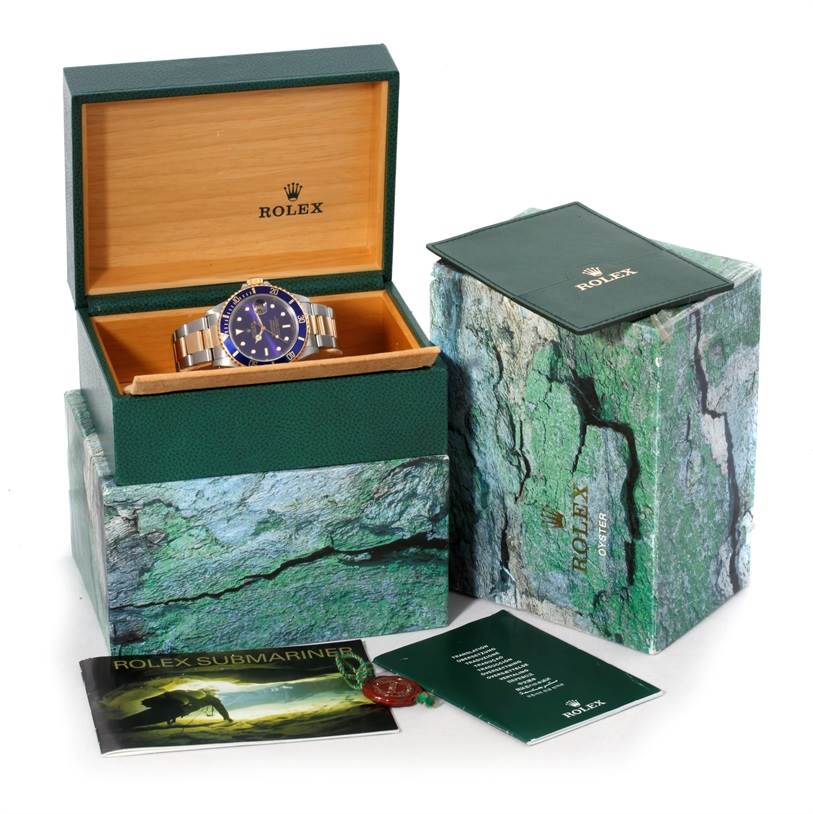 The image shows a Rolex Submariner 16613 Men's Steel and Gold (two tone) Blue Dial 16613 Men's Steel and Gold (two tone) Blue Dial watch displayed in its open box, along with its outer box, booklet, and accessories.