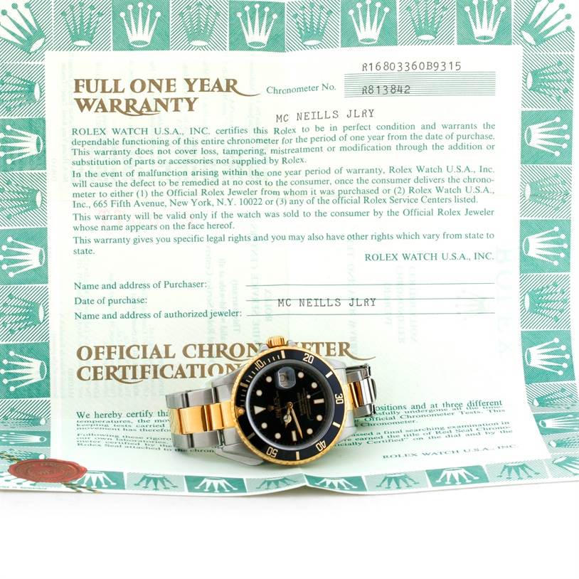 The Rolex Submariner 16803 Men's Steel and Gold (two tone) Black Dial 16803 Men's Steel and Gold (two tone) Black Dial watch is shown from a frontal angle, displayed on top of its warranty and certification papers.