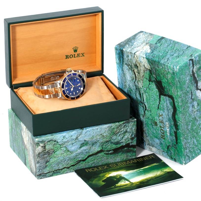 The image shows a Rolex Submariner 16613 Men's Steel and Gold (two tone) Blue Dial 16613 Men's Steel and Gold (two tone) Blue Dial watch, displayed partially side-on inside its open box with accompanying packaging and booklet.