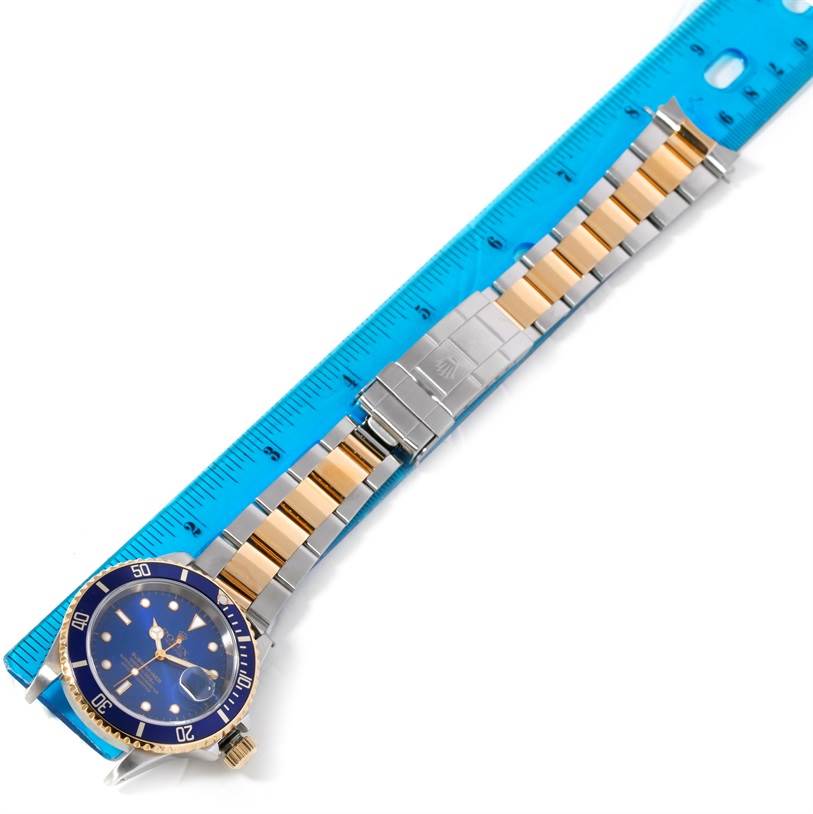 The image shows a Rolex Submariner 16613 Men's Steel and Gold (two tone) Blue Dial 16613 Men's Steel and Gold (two tone) Blue Dial watch laid flat alongside a blue ruler, displaying its blue dial, bezel, and two-tone bracelet.