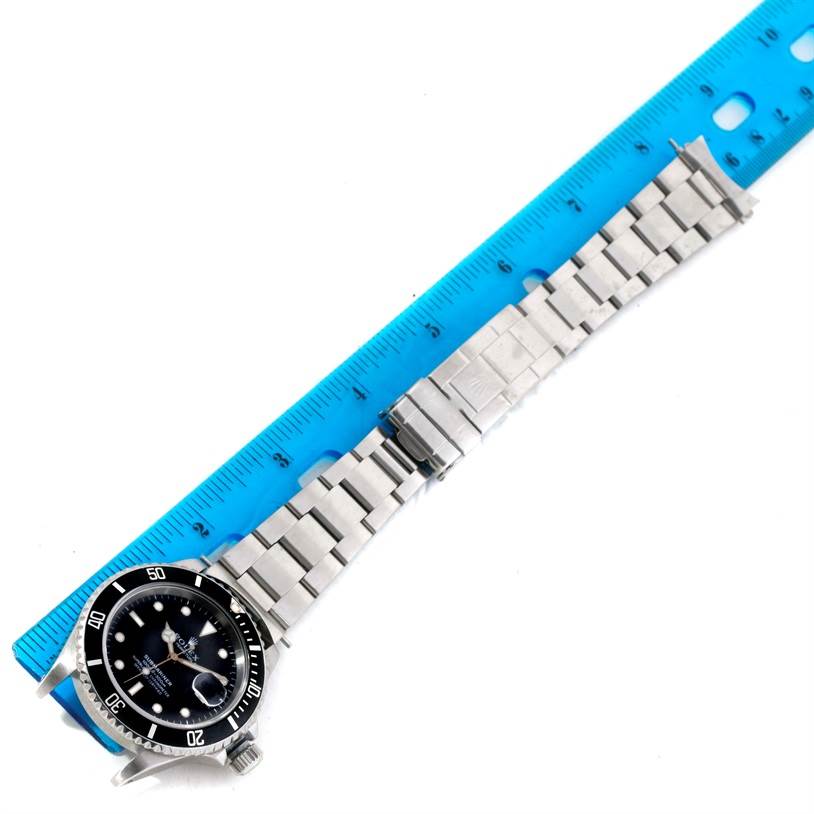 The Rolex Submariner 16610 Men
s Stainless Steel Black Dial 16610 Men
s Stainless Steel Black Dial watch is shown from a top view, displaying the face, bezel, bracelet, and clasp, next to a blue ruler.