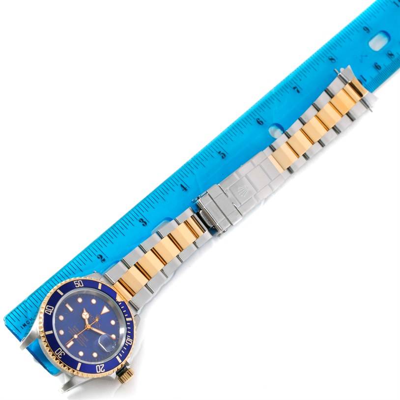 The Rolex Submariner 16613 Men's Steel and Gold (two tone) Blue Dial 16613 Men's Steel and Gold (two tone) Blue Dial watch is shown from a top angle, displaying its blue dial, bezel, and two-tone bracelet beside a blue ruler.