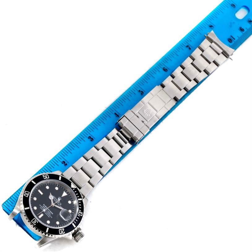 The image shows a top-down view of a Rolex Submariner 16610 Men
s Stainless Steel Black Dial 16610 Men
s Stainless Steel Black Dial watch, including the face, bezel, and linked bracelet placed alongside a blue ruler.