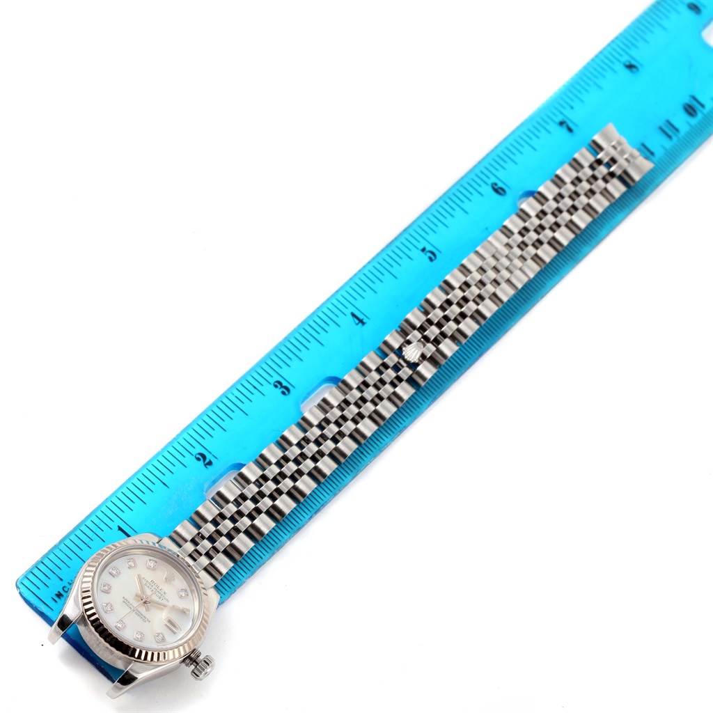 The image shows the Rolex Datejust 179174 Women
s Stainless Steel Mother of Pearl Dial 179174 Women
s Stainless Steel Mother of Pearl Dial watch face and bracelet laid next to a blue ruler measuring its length.