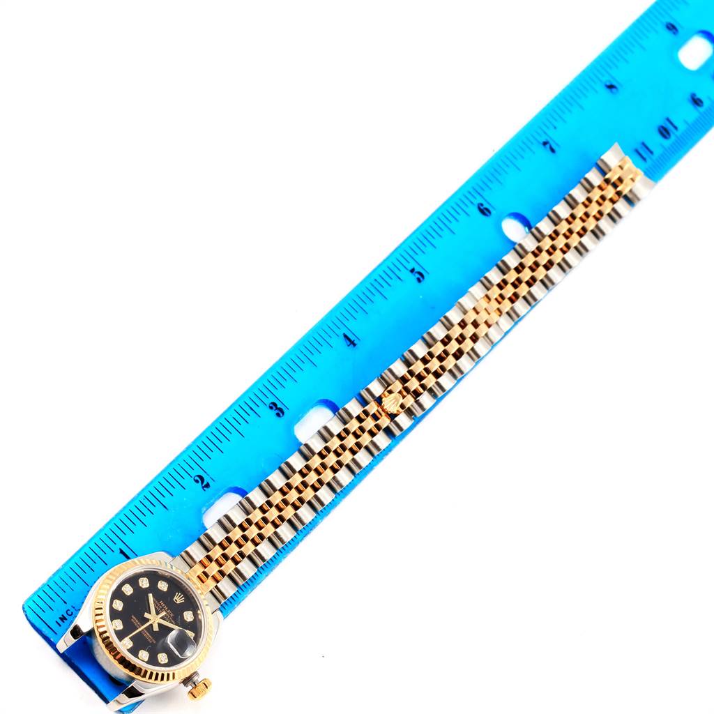The image shows a gold Rolex Datejust 179173 Women
s Steel and Gold (two tone) Black Dial 179173 Women
s Steel and Gold (two tone) Black Dial watch laid next to a blue ruler, displaying its bracelet and part of the dial.