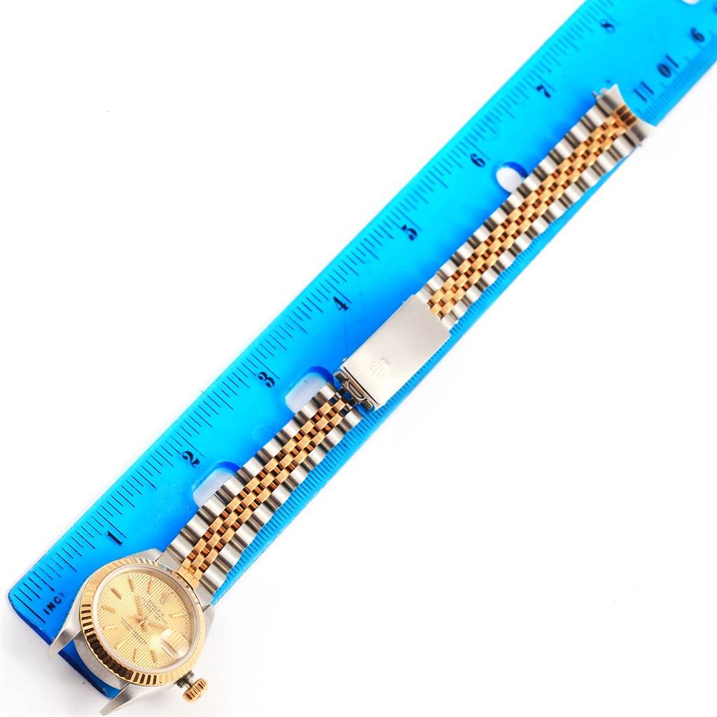 The image shows a Rolex Datejust 69173 Women's Stainless Steel Champagne Dial 69173 Women's Stainless Steel Champagne Dial watch from above, featuring its gold face and two-tone bracelet with a clasp, alongside a blue ruler.