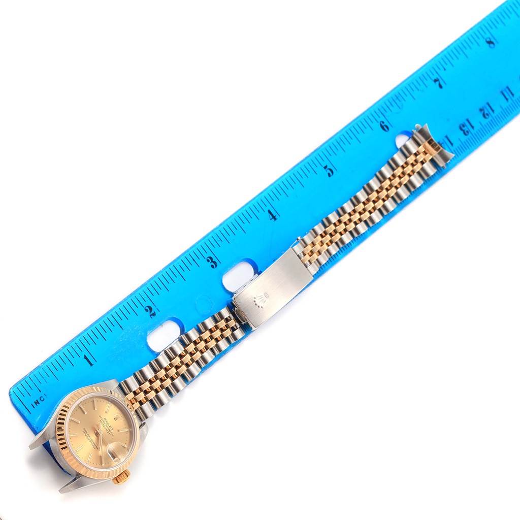 The image shows a Rolex Datejust 79173 Women's Steel and Gold (two tone) Champagne Dial 79173 Women's Steel and Gold (two tone) Champagne Dial watch laid flat beside a blue ruler, highlighting its face, band, and clasp.