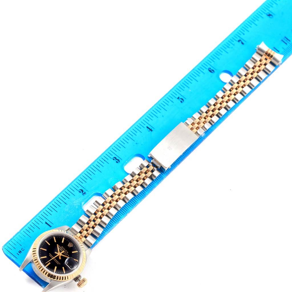 This image shows a Rolex Datejust 79173 Women's Steel and Gold (two tone) Black Dial 79173 Women's Steel and Gold (two tone) Black Dial watch with its black dial, bezel, and jubilee bracelet laid next to a ruler for scale.