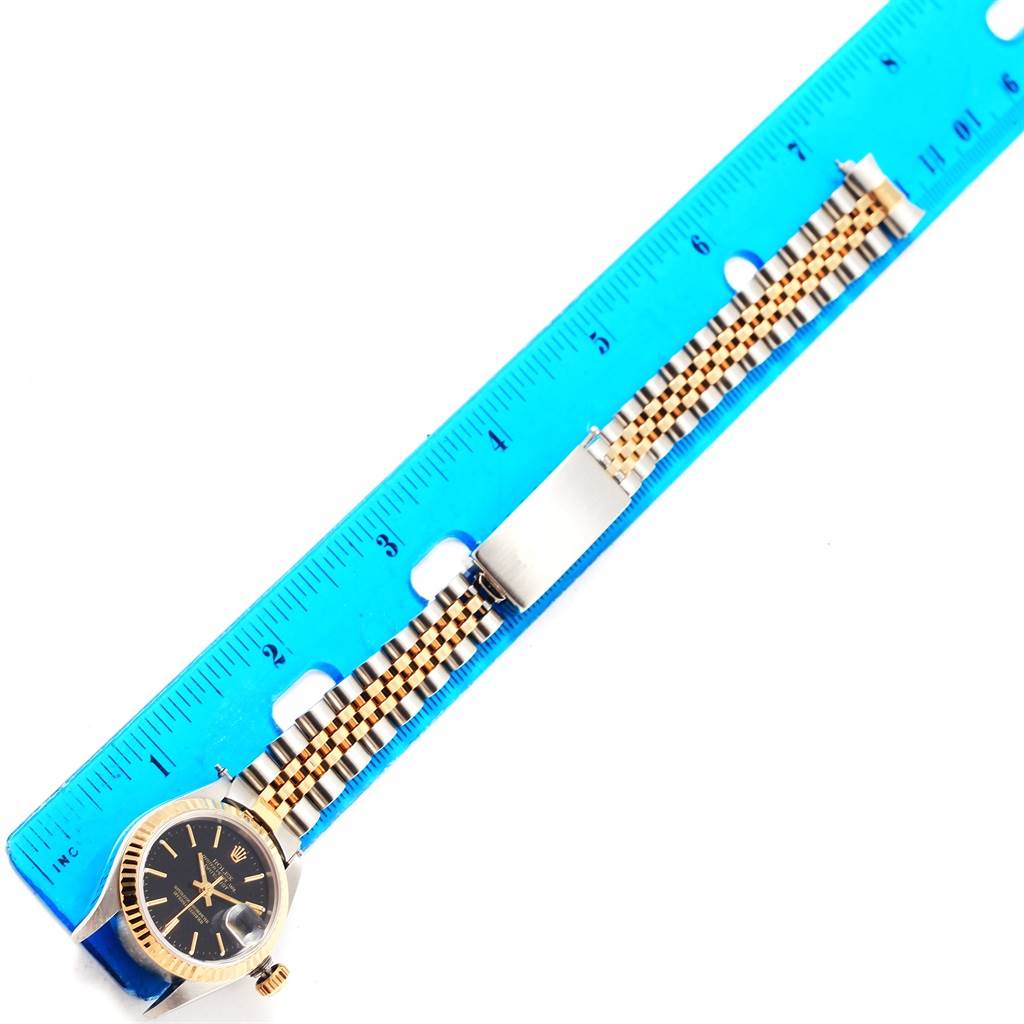 The Rolex Datejust 79173 Women
s Steel and Gold (two tone) Black Dial 79173 Women
s Steel and Gold (two tone) Black Dial watch is shown horizontally, displaying the face, crown, and detailed bracelet with measurement from a blue ruler.