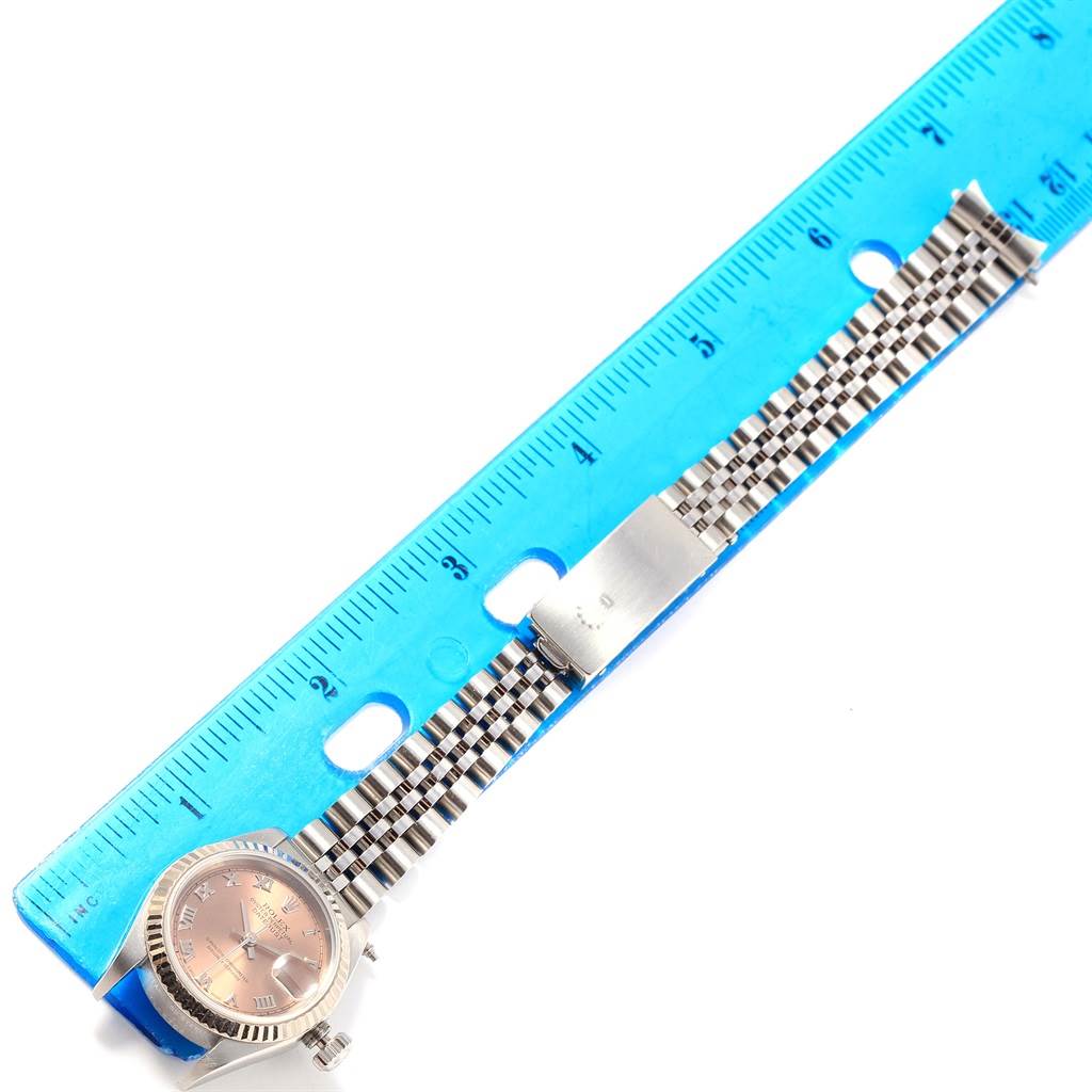 The image shows a Rolex Datejust 79174 Women
s Stainless Steel Pink Dial 79174 Women
s Stainless Steel Pink Dial watch with a close-up of its face, crown, and Jubilee bracelet, measured against a blue ruler.