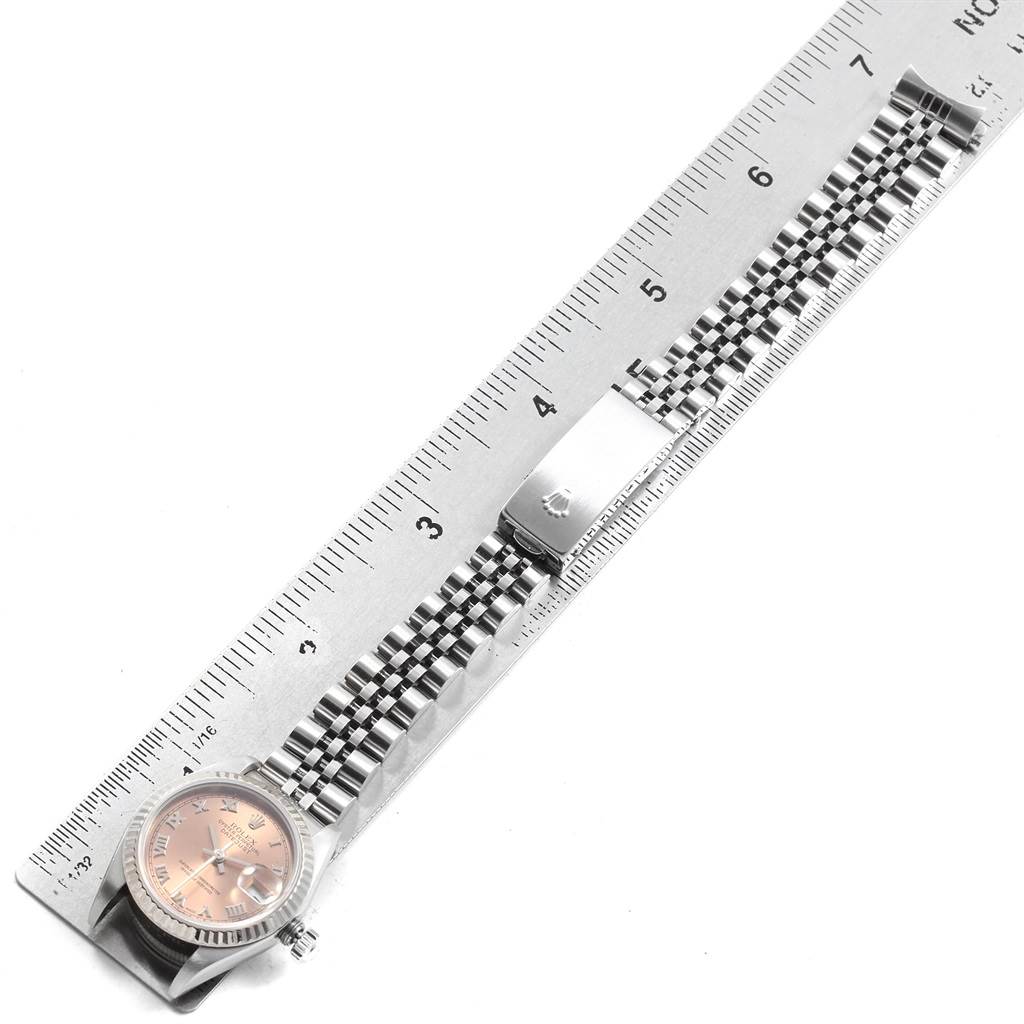 The image shows a Rolex Datejust 79174 Women
s Stainless Steel Pink Dial 79174 Women
s Stainless Steel Pink Dial watch laid flat next to a ruler, highlighting its dial, crown, and Jubilee bracelet with clasp.