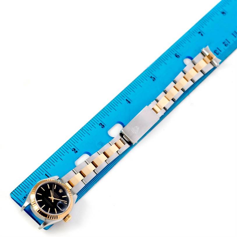 The Rolex Datejust 79163 Women's Steel and Gold (two tone) Black Dial 79163 Women's Steel and Gold (two tone) Black Dial watch is displayed flat next to a blue ruler, showing the dial, bracelet, and clasp.