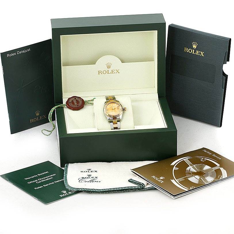 The Rolex Datejust 179173 Women's Steel and Gold (two tone) Champagne Dial 179173 Women's Steel and Gold (two tone) Champagne Dial watch is displayed inside an open box with accompanying booklets and accessories surrounding the box.