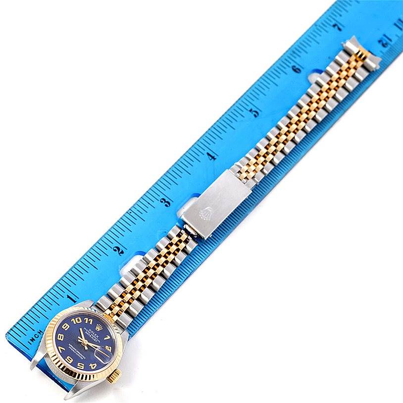 The Rolex Datejust 69173 Women
s Stainless Steel Blue Dial 69173 Women
s Stainless Steel Blue Dial watch is shown laid flat with a blue face, gold bezel, and Jubilee bracelet next to a blue ruler.