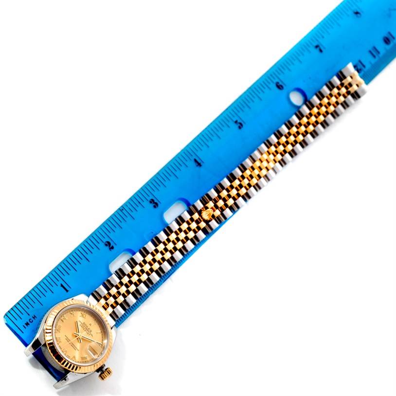 The image shows a Rolex Datejust 179173 Women's Steel and Gold (two tone) Champagne Dial 179173 Women's Steel and Gold (two tone) Champagne Dial watch laid flat alongside a blue ruler, highlighting the watch's face and bracelet.