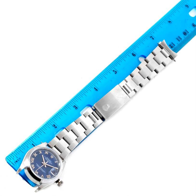 The image shows a Rolex Mid-Size 68240 Women
s Stainless Steel Blue Dial 68240 Women
s Stainless Steel Blue Dial model watch laid flat against a blue ruler, displaying its silver bracelet and blue dial.