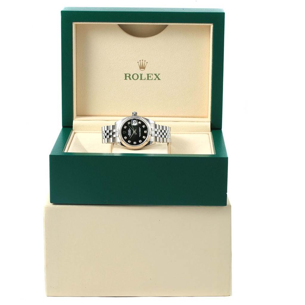 The image shows a front view of a mid-size Rolex watch with a black dial and a metal bracelet in an open green Rolex box.