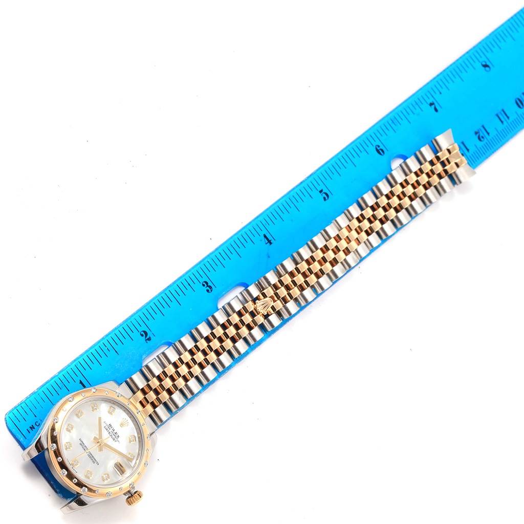This image shows a Mid-Size Rolex watch laid flat alongside a blue ruler, highlighting the case and bracelet.