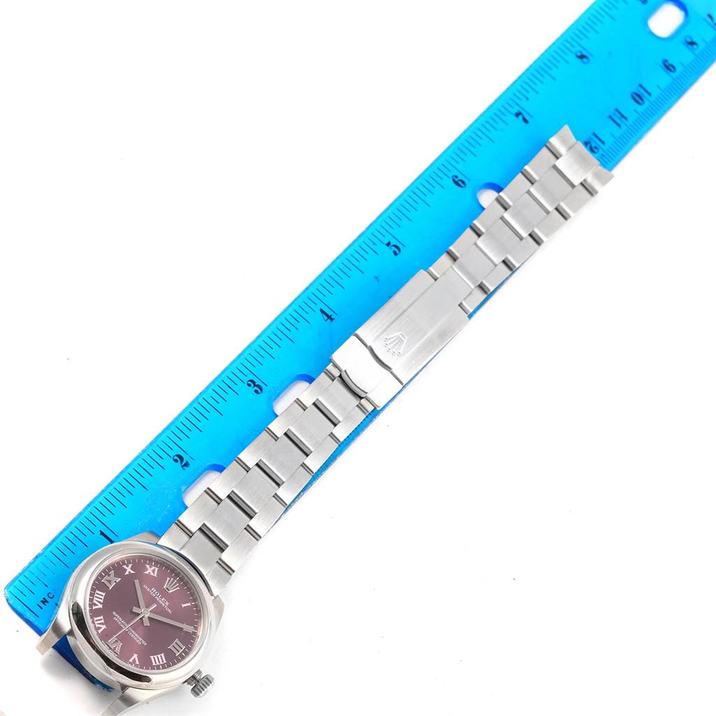 The image shows a Mid-Size Rolex watch with a maroon dial and stainless steel bracelet, laid flat next to a blue ruler.